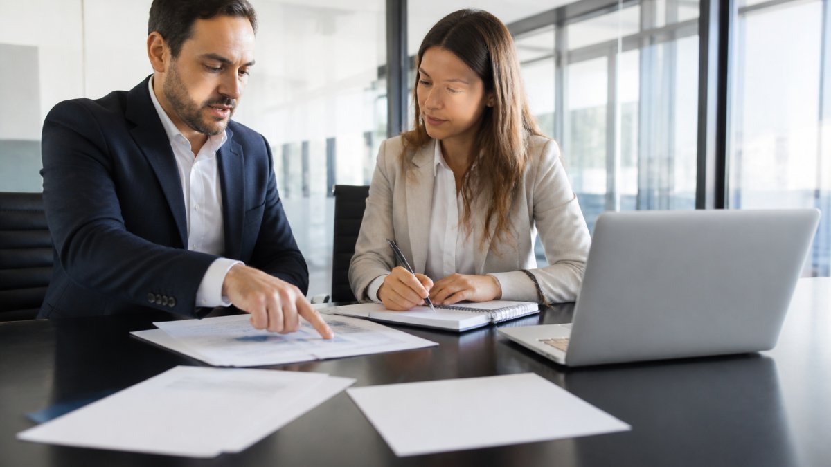 Two business professionals reviewing printed strategy documents and a laptop at a conference table in a bright office, one pointing at a page while the other takes notes